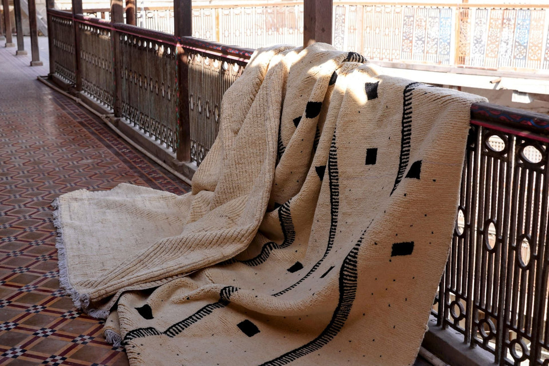 Thick-pile cream rug with black geometric marks draped over a carved wooden balcony rail in daylight.