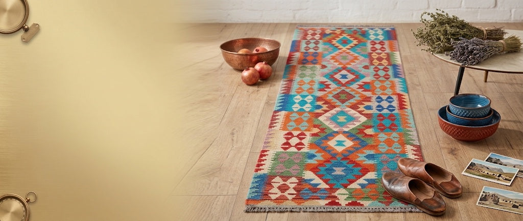 Orange, red and turquoise diamond-pattern kilim runner on a light oak floor beside a copper bowl of pomegranates and leather slippers
